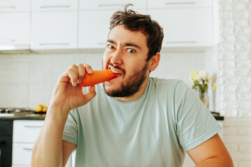 Front view of a young man biting a raw carrot with a ferocious and unhappy expression. Healthy...