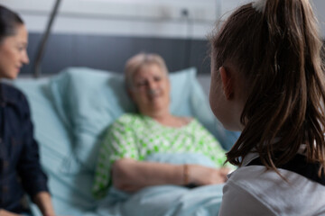 Elderly woman out focus, lying in hospital bed chatting with unidentified little girl. Family...