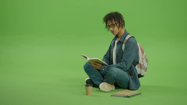 Young Multicultural Man in Glasses and Denim Shirt Looking to Book,Turn Pages of Book and Smiling. Portrait of Handsome Guy Sitting with Books,Notes and Coffee on Green Screen Background