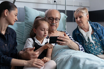 Little girl showing cell phone videos to grandfather hospitalized in geriatric clinic. Elderly man...