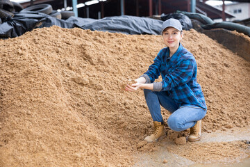 Woman farmer squatting at large heap of brewer's spent grain and holding bunch in hands.