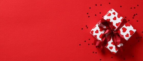 Gift box with red ribbon bow and hearts shaped confetti on red background. Happy Valentines Day banner design, header template. Top view with copy space.
