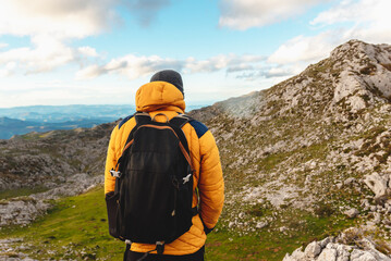 man from behind with backpack and warm clothes, with yellow mountain jacket observing the mountain peak that he is going to climb. climbing mountain peaks