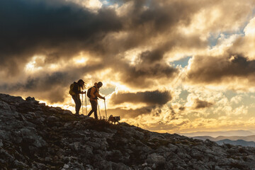 two hikers with their dog in silhouette descending a mountain at sunset on a cloudy day. couple doing trekking. sport and outdoor adventure.
