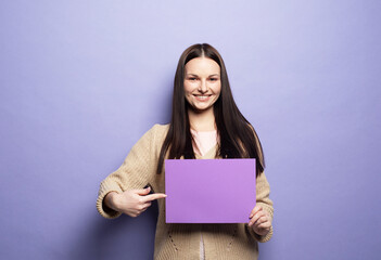 Fototapeta premium Young woman holding purple blank advertising board standing on lilac background in studio