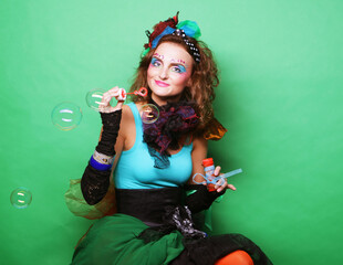 Young curly woman with creative make-up blowing soap bubbles over green background.