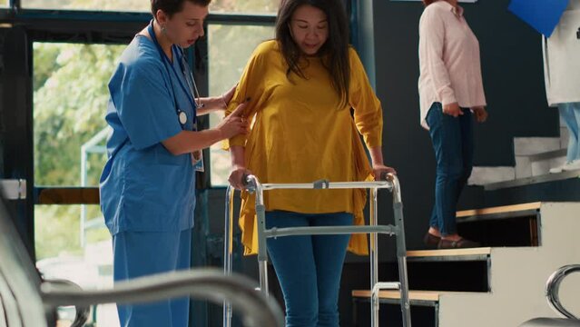 Nurse helping asian woman to walk with walking frame, suffering from physical injury and impairment. Injured patient with leg pain recovering after fracture and trauma. Handheld shot.