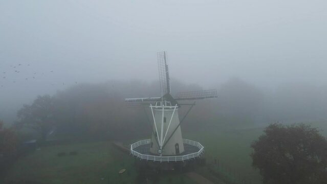 Drone puling back from beautiful old windmill in a fog covered landscape with a flock of birds flying over treeline