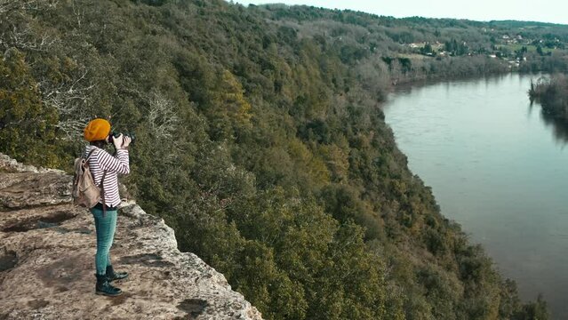 Aerial photo of a woman on the edge of a cliff taking landscape photos by a river, she is wearing an orange cap, long traveling forward