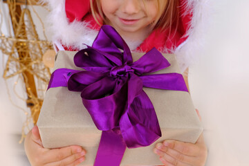 gift with purple ribbon ​​in hands of toddler, small child, blonde girl 3 years old in red New Year's dress close-up, gift from Santa Claus for Christmas, happy childhood, dream of fairy tale, magic