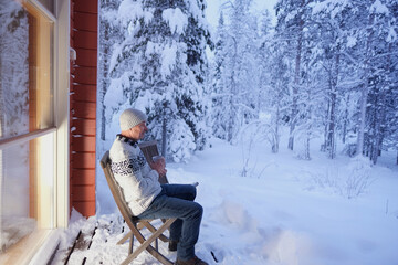positive mature man, senior 60 years sit on cozy wooden chair on terrace of country house in winter forest and drink hot tea, early evening, warm light from large window, enjoying winter nature