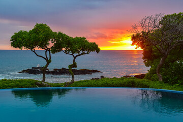 sunset over ocean with infinity pool and tropical trees and volcanic rocks with crashing waves