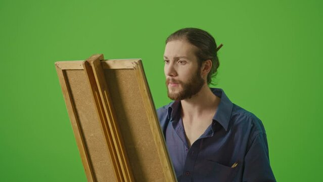 Young Artist in Denim Shirt and Apron Working on Creation of New Painting. Motivated Bearded Man Draws with Brush on Canvas on Easel on Green Screen,Chroma Key. The Creativity And Art Concept