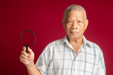 Senior man holding a magnifying glass and looking at the camera while standing on a red background