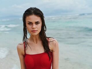 Beautiful woman on the beach by the sea in a red swimsuit looks at the camera