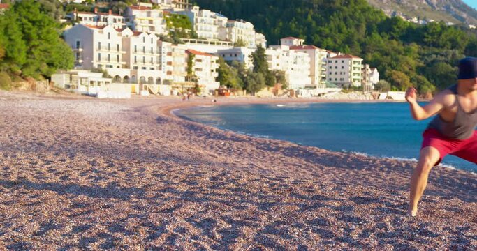 Barefoot man in Panama shorts runs along beach towards sea with a funny crab gait, two dogs are chasing him. Family rest vacation parent kids having fun fooling around on the beach active games pet