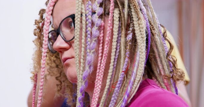 Young blonde woman with glasses is sitting and waiting for master to unwind or braid her multicolored box braids, side view, close up. Different female hairstyles for long hair.