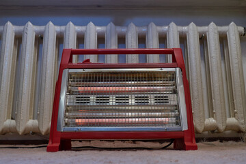 The red heater lies on the carpet in the bedroom. In the background is a cast iron radiator. The house has poor heating, you have to connect a working heating element, an additional source of heat.