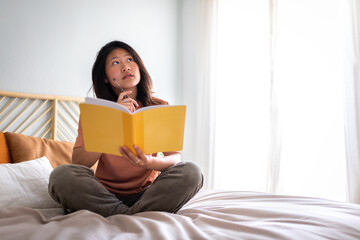 Teen asian girl sitting on bed thinking, using diary and pen to write down thoughts and ideas. Copy...
