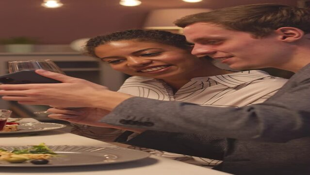 Vertical shot of smiling young Caucasian man showing photos on his smartphone to his Biracial female friend, sitting at festive dinner table, drinking wine and chatting