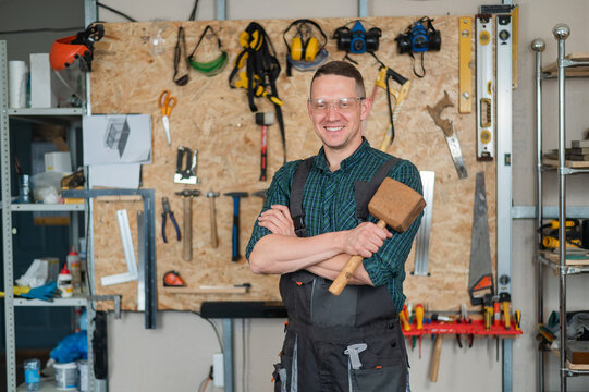 Portrait of a carpenter in goggles and overalls holding a wooden hammer in the workshop against the background of a wall with tools.