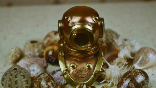 A copy of an old copper diving helmet surrounded by beautiful multi-colored sea shells against the background of the table surface imitating sea sand. Marine romance concept