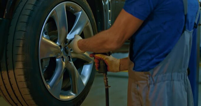 Mechanic in blue overalls is unscrewing lug nuts with a pneumatic impact wrench. Specialists removes the wheel in order to fix a component on a vehicle.
