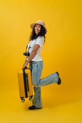 Happy Traveller curly latin woman wears white t-shirt with suitcase bag and retro camera isolated on yellow background.