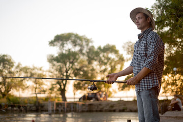 Man fishing outside in evening on lake in summer, enjoy spending time in nature, side view portrait of male in casual checkered shirt