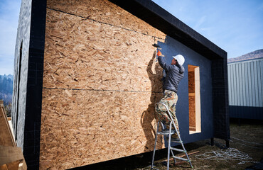 Male painter using paint roller, doing exterior paint work in a black color, standing on a ladder. Man worker building wooden frame house. Carpentry and construction concept.