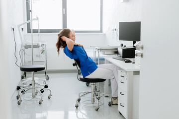 Young woman doctor stretching at hospital office desk.