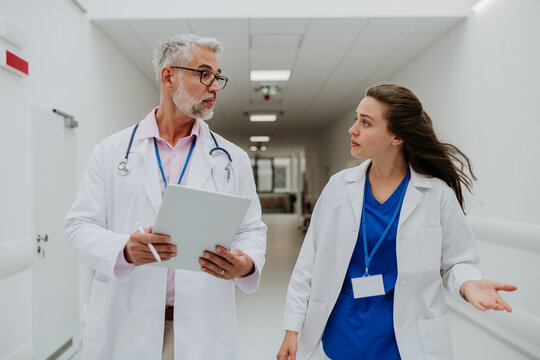 Older Doctor Giving Advise To His Younger Colleague, Discussing At Hospital Corridor. Health Care Concept.