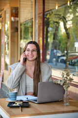 Smiling woman working with a laptop and talking on phone in a coffee shop. Digital nomad.