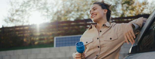 Young woman holding power supply cable from her electric car, prepared for charging it in home,...