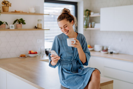 Young woman with smartphone enjoying cup of coffee at morning, in her kitchen.