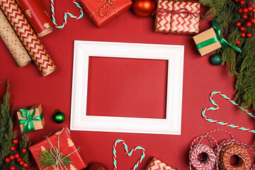 Blank white photo frame surrounded by Christmas gifts, wrapping paper and juniper branches on red background.