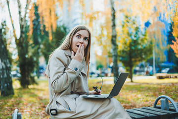 A cute fair-haired young woman is sitting in an autumn park on a bench and working on a laptop. Remote work
