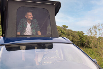 smiling young man looking out over the landscape sitting in the roof tent of his camper van,...