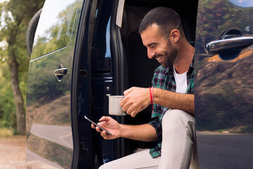 smiling young man using his mobile phone sitting in the doorway of his camper van while enjoying a...