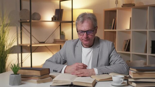 Overload information. Tired aged man. Lecture preparing. Exhausted smart male taking off spectacles falling asleep on pile of books in light shadow room interior.