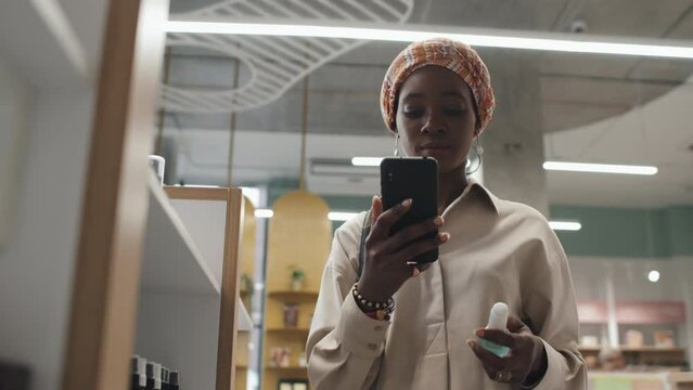 Young black woman reading online information about new skincare product in mobile phone while standing by display with beautycare items in cosmetic supermarket