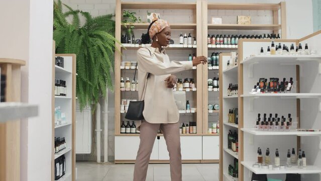 Young black woman with shopping basket walking along displays with beautycare products in large modern cosmetic supermarket and taking one of skincare items
