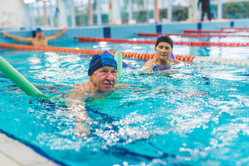Proud involved caucasian senior man in a swimming cap using bright green pool noodle to help him during swimming. Active leisure time. Blurred caucasian woman behind him. High quality photo