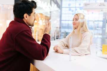 Back view from shoulder of handsome man chatting through glass partition with charming young blonde woman standing in bright hall of shopping mall. Concept of lifestyle social distancing and work.