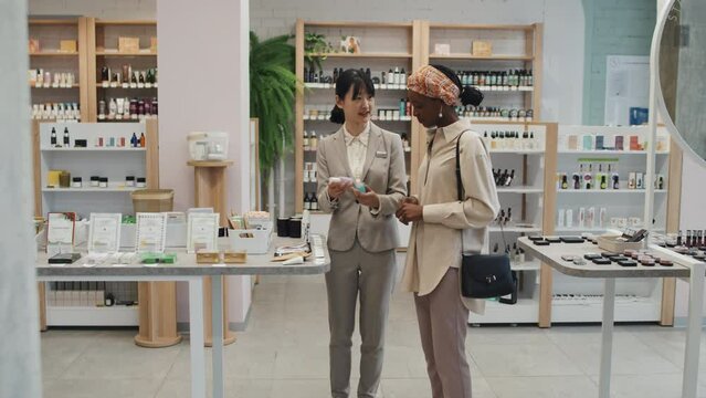 Young shop assistant and consumer walking along displays in cosmetic supermarket and stopping by table with new collection of skincare products to discuss and compare their characteristics
