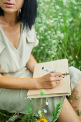 Female woman with pen writing or painting, handwriting on notebook on flower blooming meadow