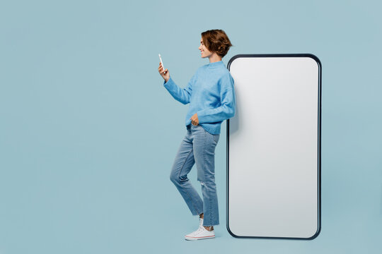 Full body happy side view young woman wear knitted sweater near big huge blank screen mobile cell phone with workspace mockup area use smartphone isolated on plain pastel light blue cyan background.