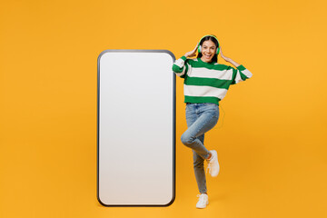 Full body young latin woman wear casual green knitted sweater headphones big huge blank screen mobile cell phone smartphone with area listen music isolated on plain yellow background studio portrait.