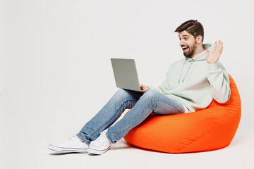 Full body young smiling fun IT man wears mint hoody sit in bag chair hold use work on laptop pc...