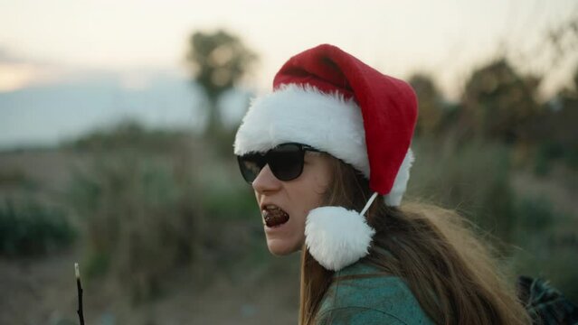 A girl in a red Santa hat eats fried marshmallows on the beach by the sea and wants to spit it out, it's hot.